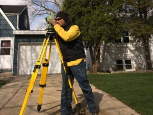 A surveyor in a yellow jacket adjusting a theodolite on a bright yellow tripod in a suburban yard.