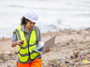 Field surveyor wearing safety gear using a laptop and handheld surveying device on a construction site to collect topographic data for drainage and grading analysis.